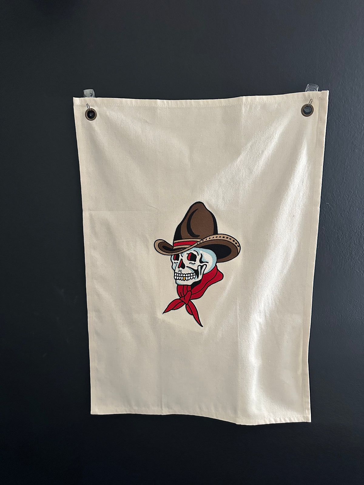 An off-white canvas flag with two metal grommets on the left side, featuring a central graphic of a skull wearing a brown cowboy hat and a red bandana, displayed against a dark gray wall.