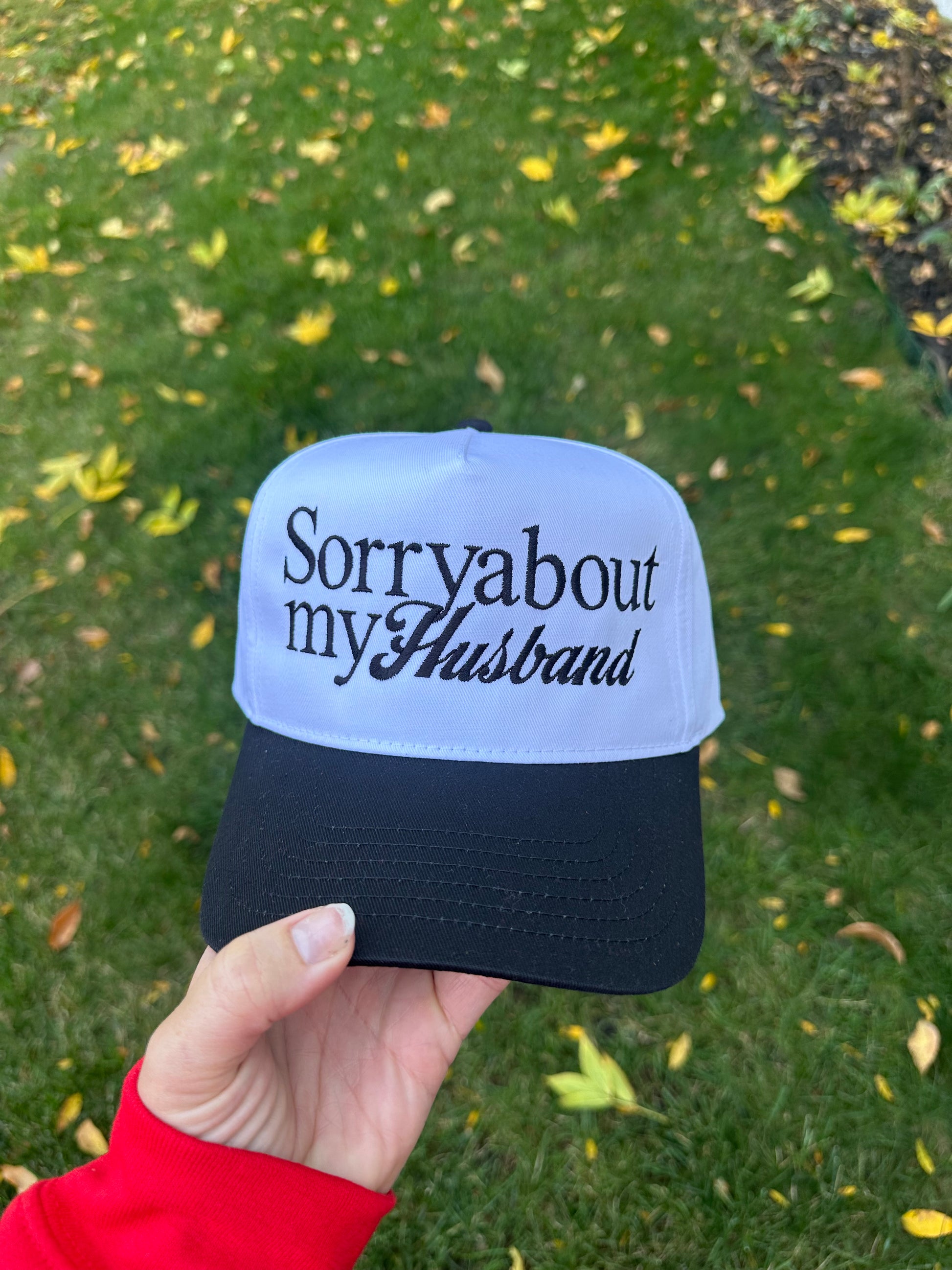 Cap with 'Sorry about my husband' text held by a hand against a grassy background