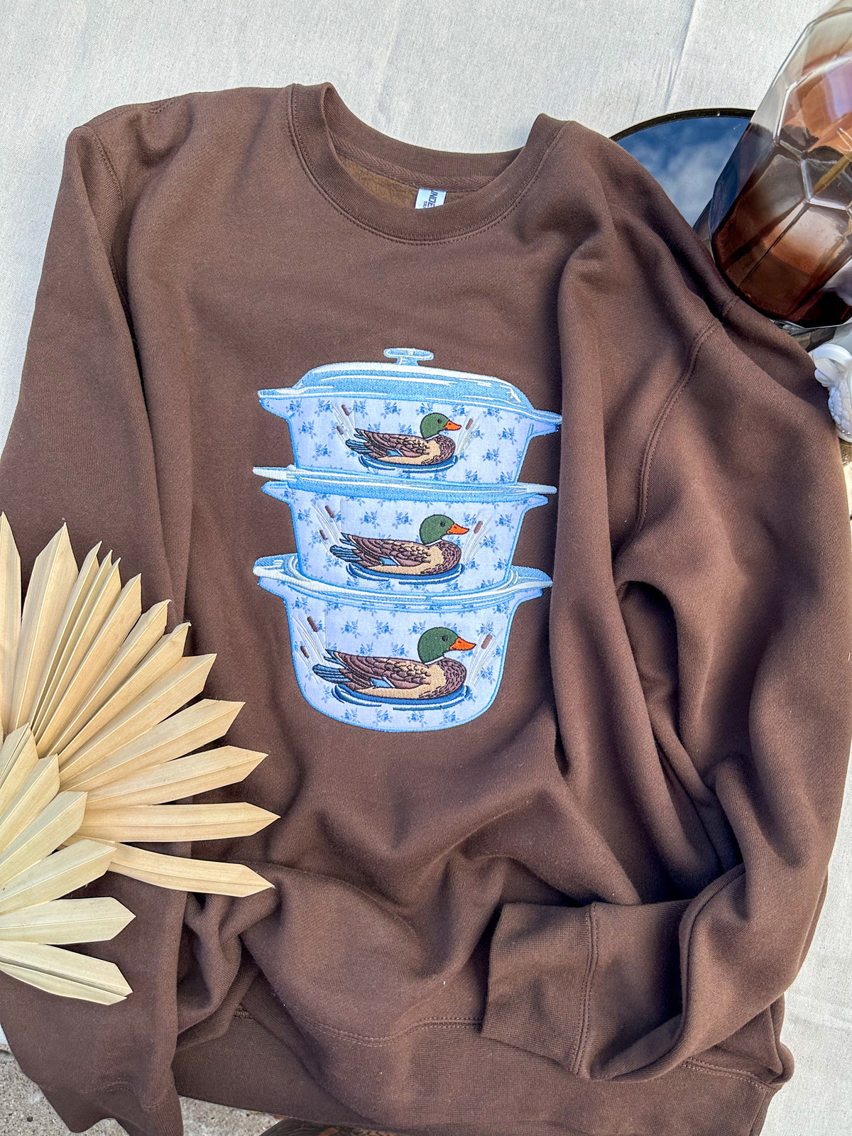 A brown crewneck sweatshirt featuring a graphic of three stacked blue ceramic Dutch ovens with mallards, styled flat-lay with a dried palm leaf and a glass bottle on a light stone background.