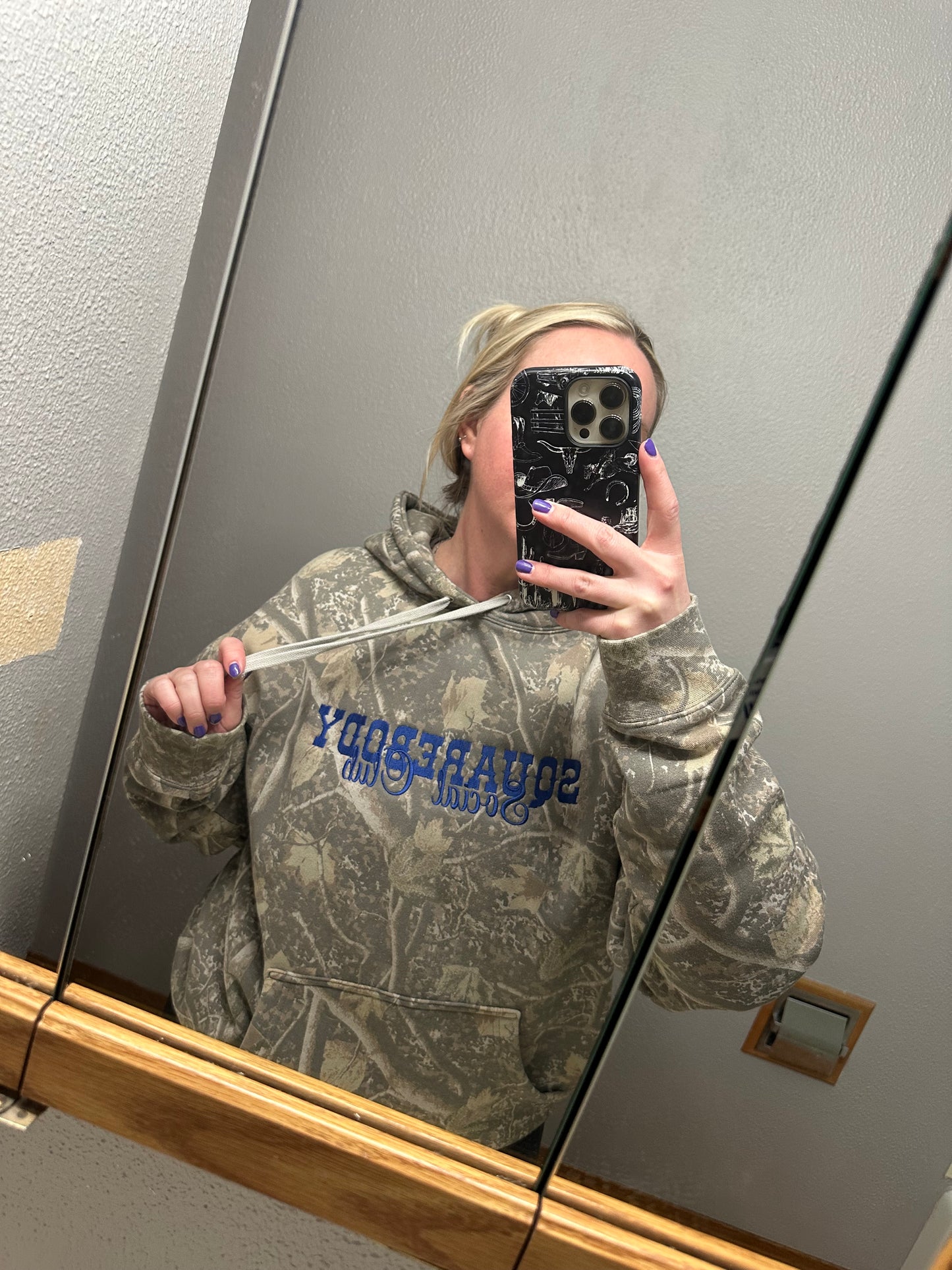 Person wearing a camouflage hoodie with squarebody social club
, taking a mirror selfie.