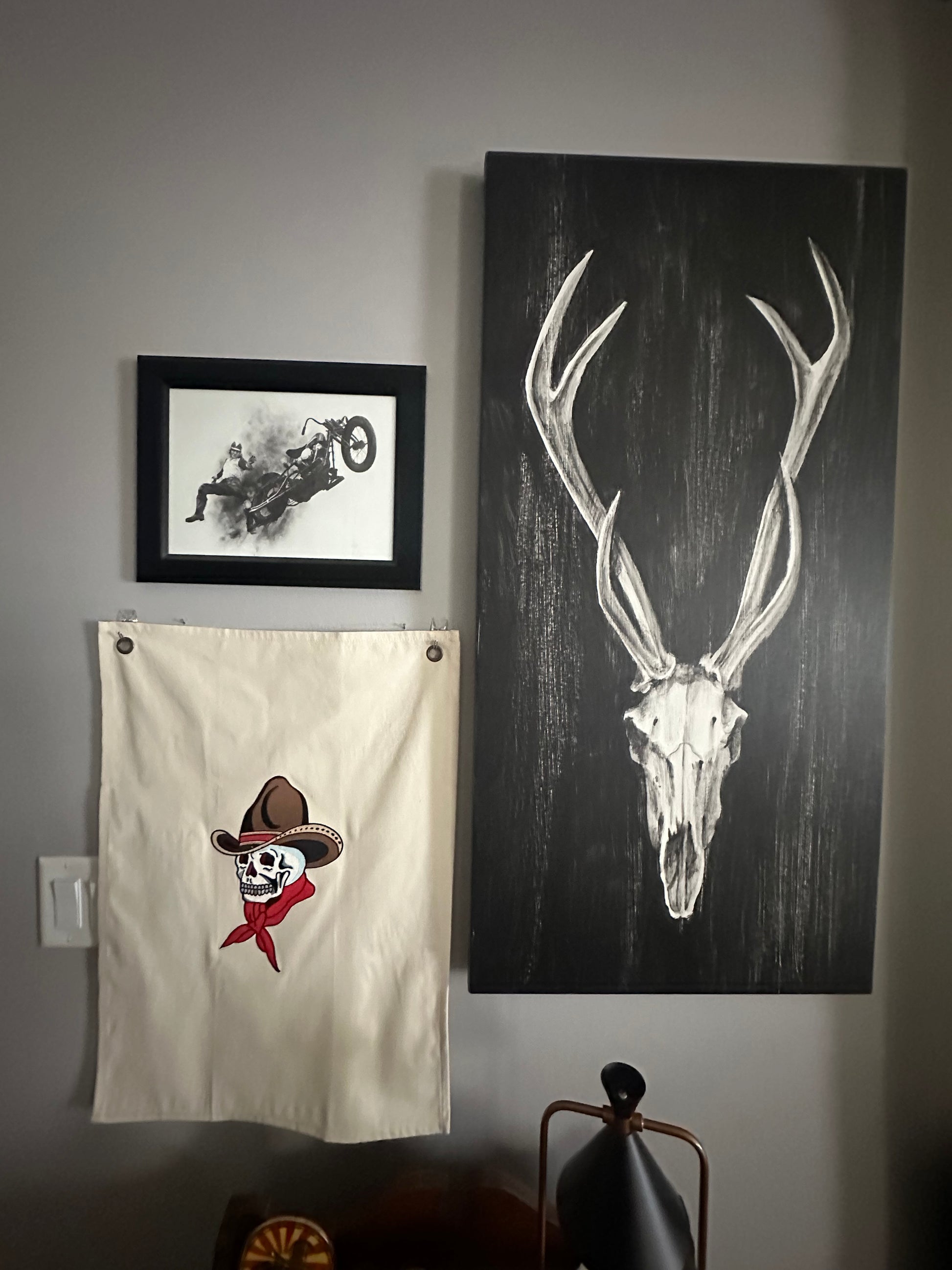 A wall-mounted display featuring a large rectangular black art print with a white antler skull design, a small black-framed black-and-white print, and an off-white flag with a pirate skull wearing a brown hat and red bandana. A lamp is partially visible on the right against a gray wall.