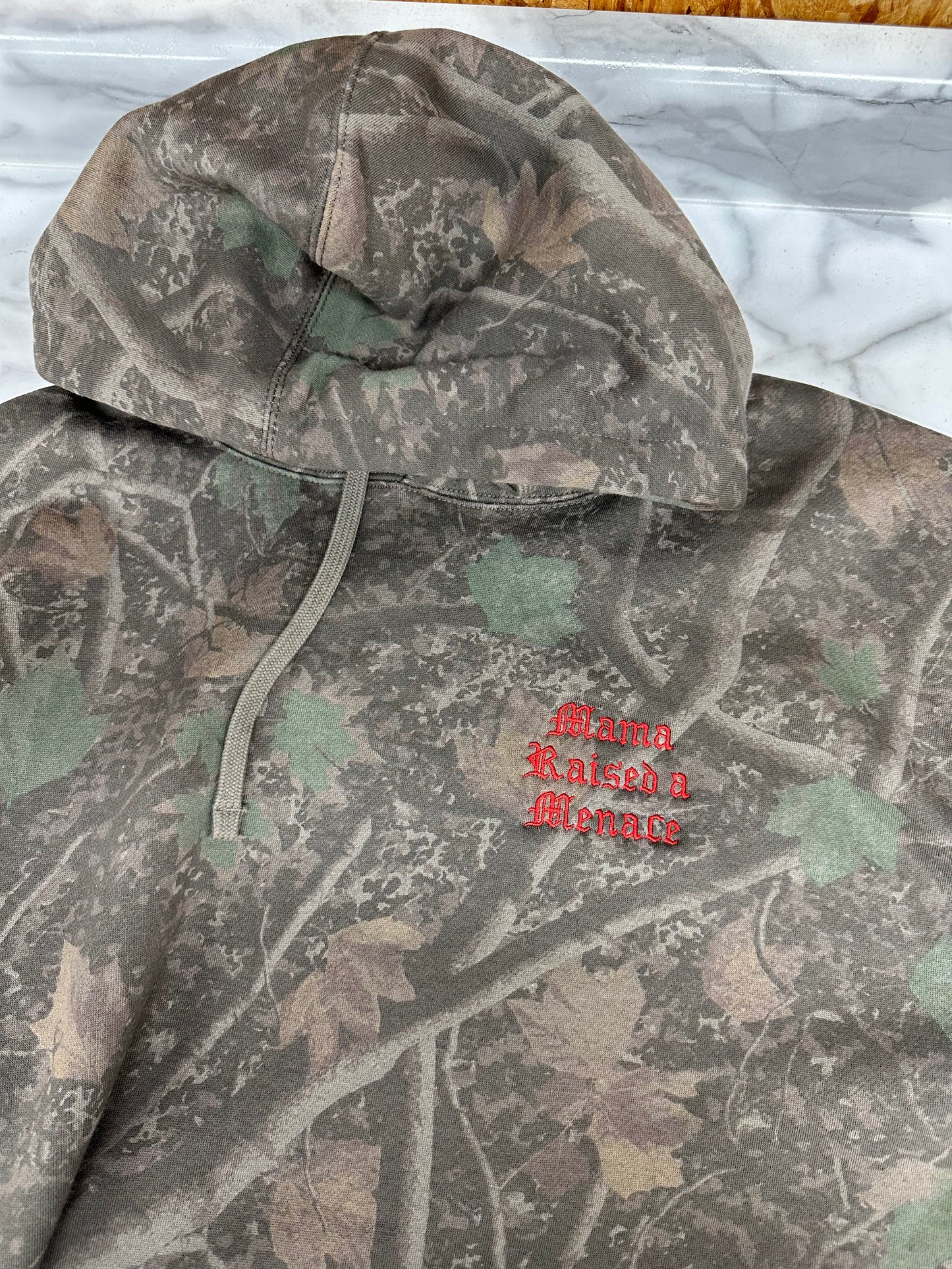 A camouflage hooded sweatshirt laid flat, showing a woodland tree-branch and leaf pattern in gray, brown, and green, with a small red embroidered logo on the chest. Mama raised a menace 