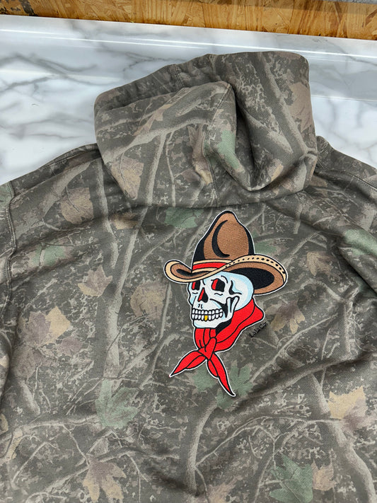 A camouflage hoodie with a skull and bandana graphic on the chest, laid flat on a white marble-patterned surface.