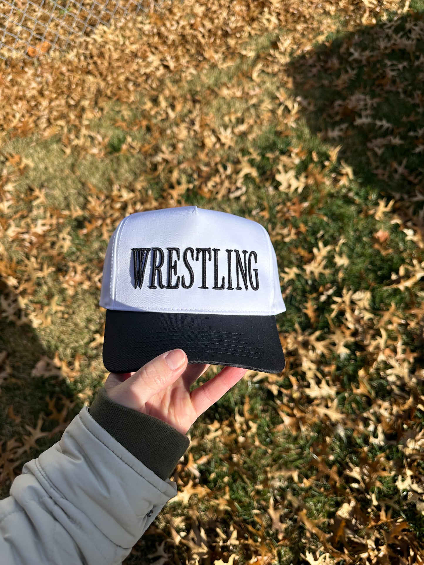 Hand holding a cap with 'Wrestling' text against a grassy background, 3d puff embroidery
