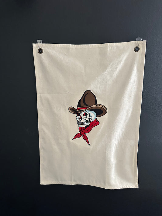 An off-white canvas flag with two metal grommets on the left side, featuring a central graphic of a skull wearing a brown cowboy hat and a red bandana, displayed against a dark gray wall.