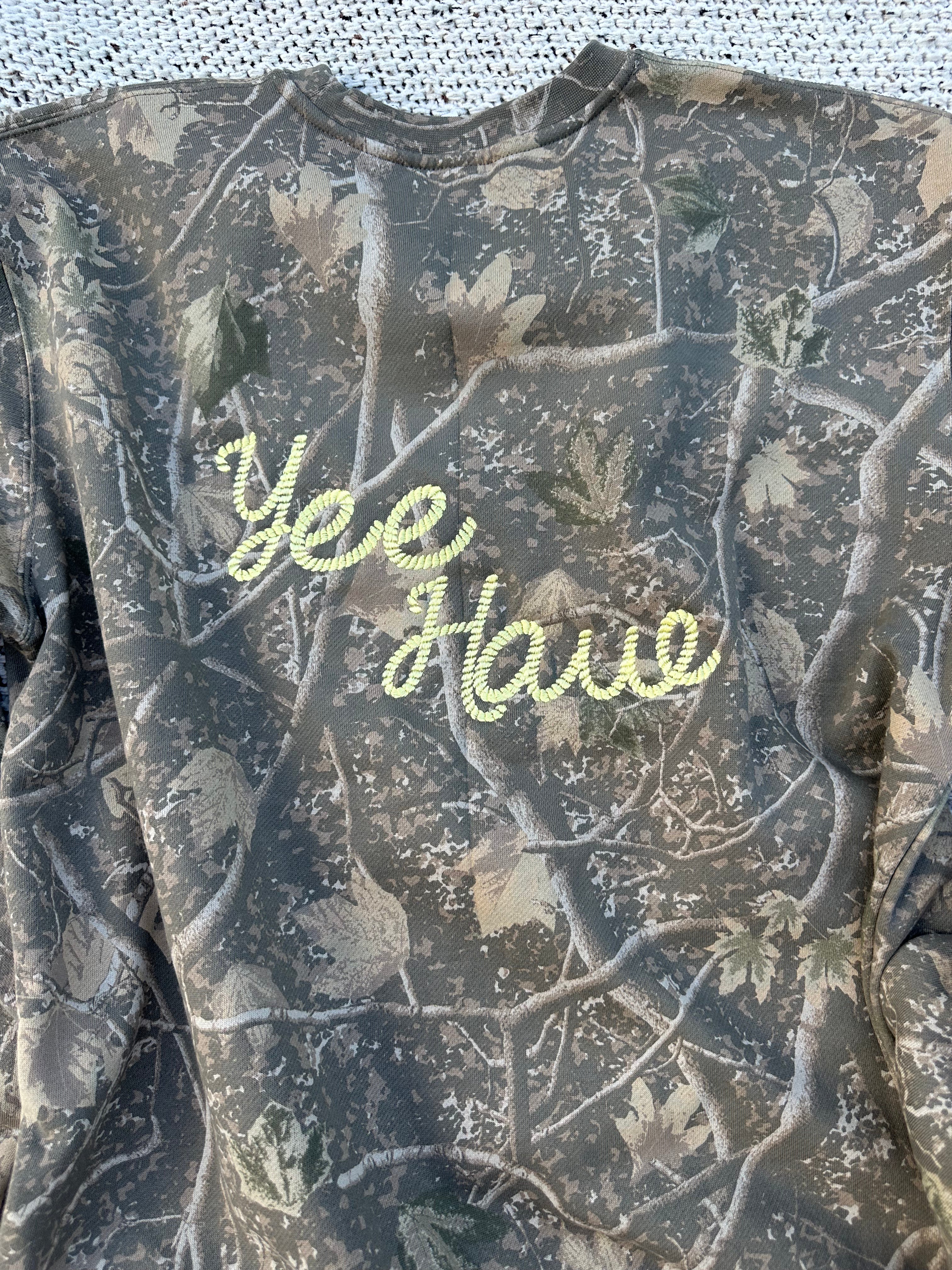 A camouflage long-sleeve shirt with 'Yee Haw!' embroidered in light green script across the chest, laid flat on a white textured surface.