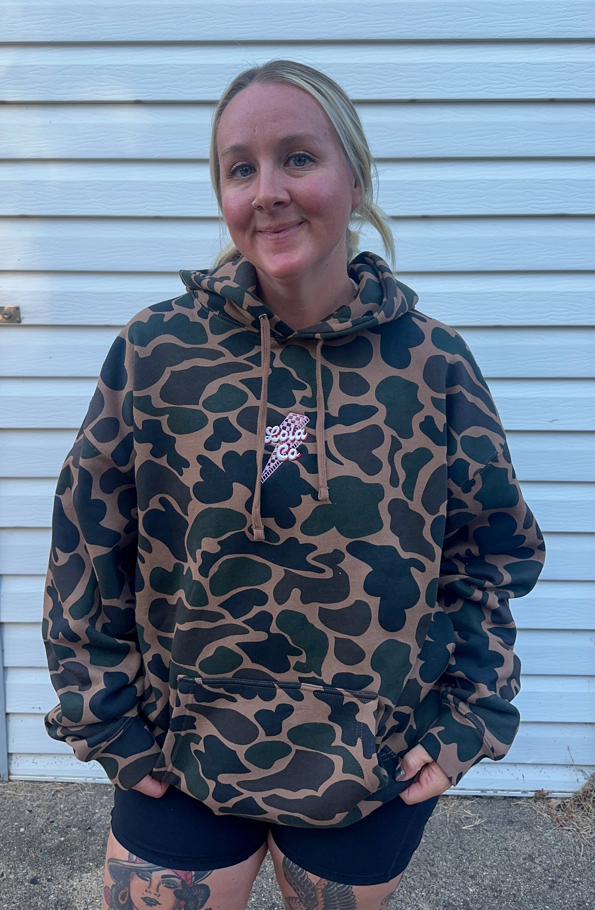 Person wearing a camouflage hoodie with a brand logo, standing against a light blue wall.