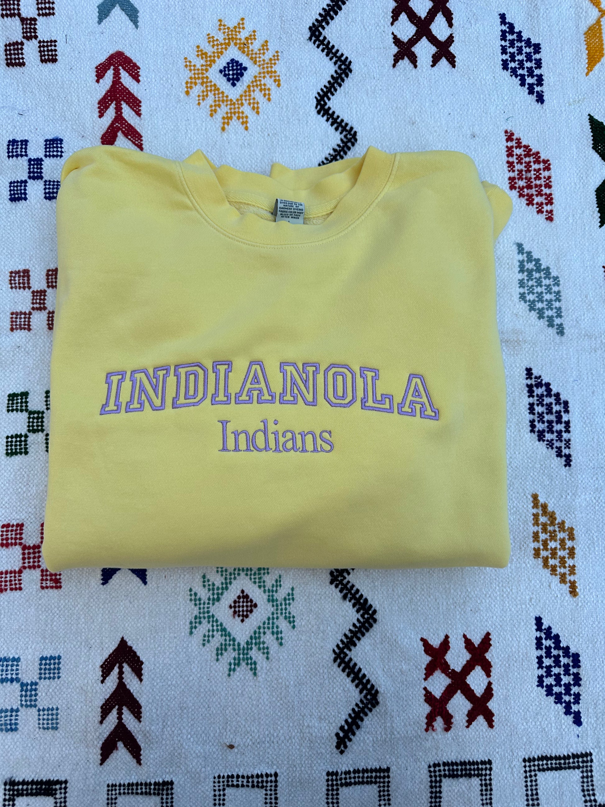 Folded yellow sweatshirt with 'INDIANOLA Indians' text on a patterned fabric background