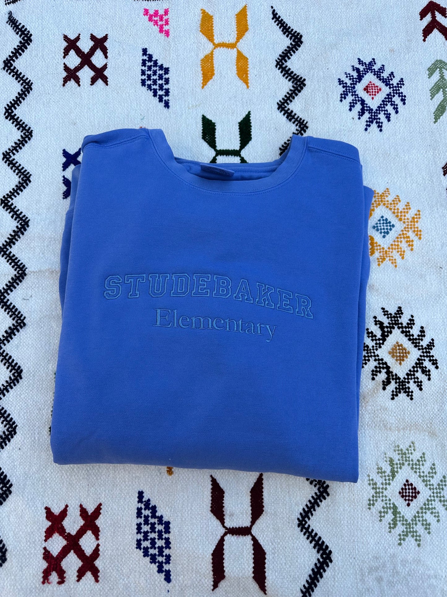 Folded blue sweatshirt with 'STUDEBAKER Elementary' text on a patterned fabric background