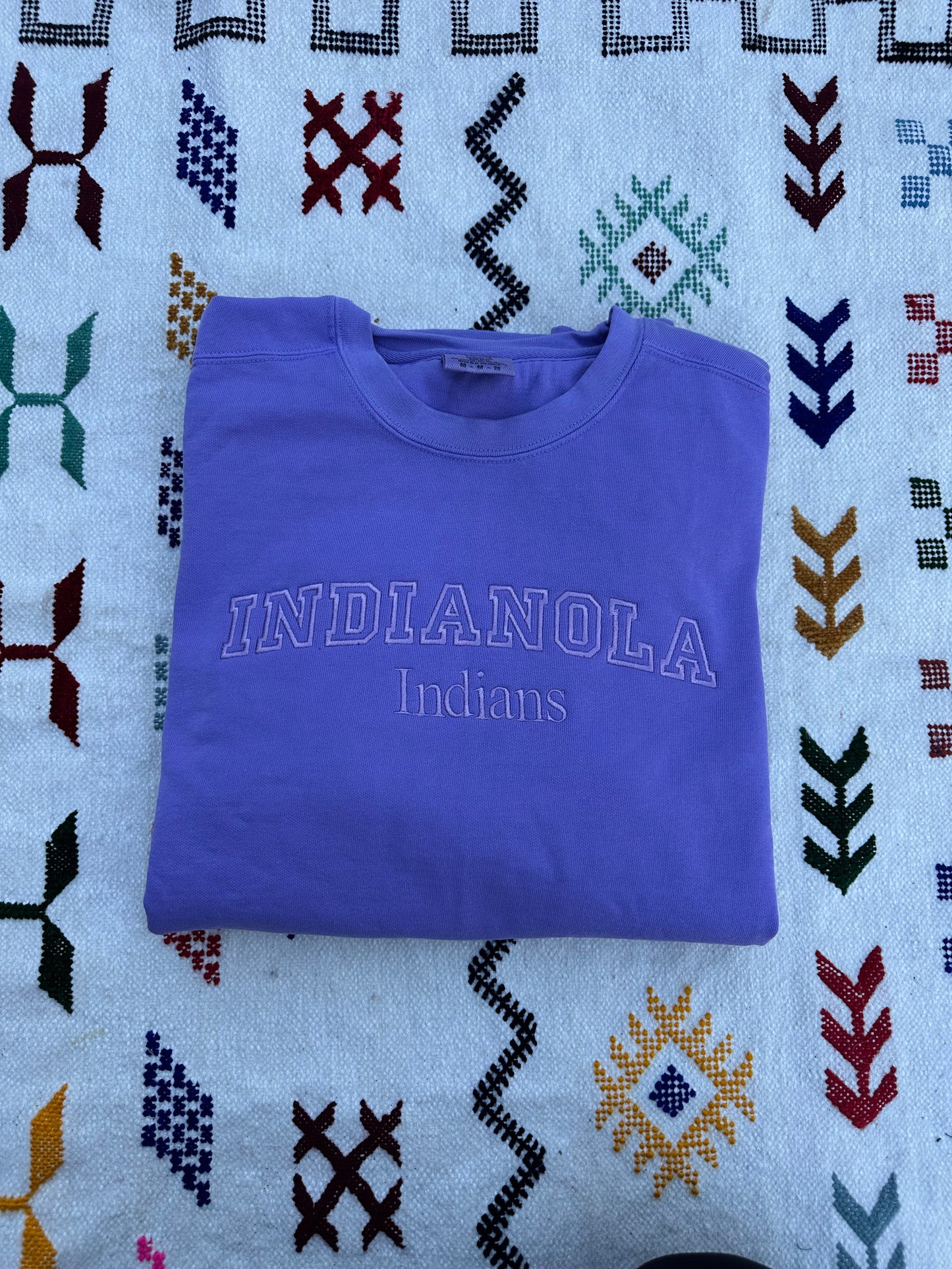 Folded purple sweatshirt with 'INDIANOLA Indians' text on a patterned fabric background