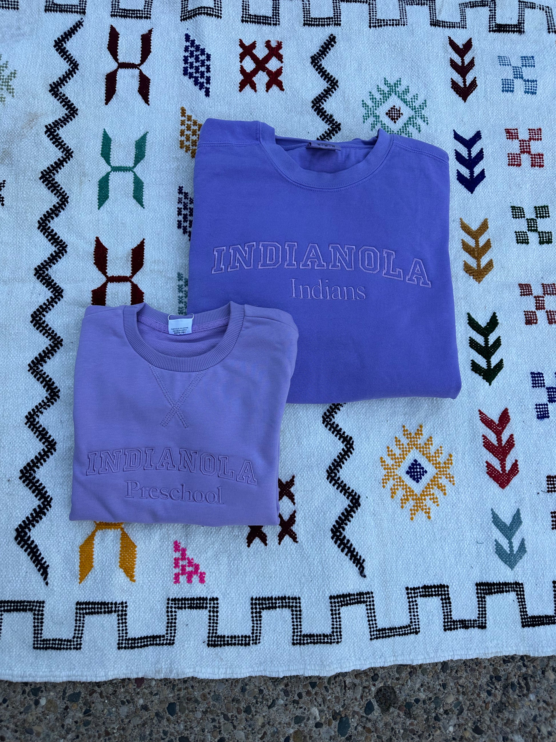 Two purple shirts with 'INDIANOLA Indians' text on a patterned fabric background