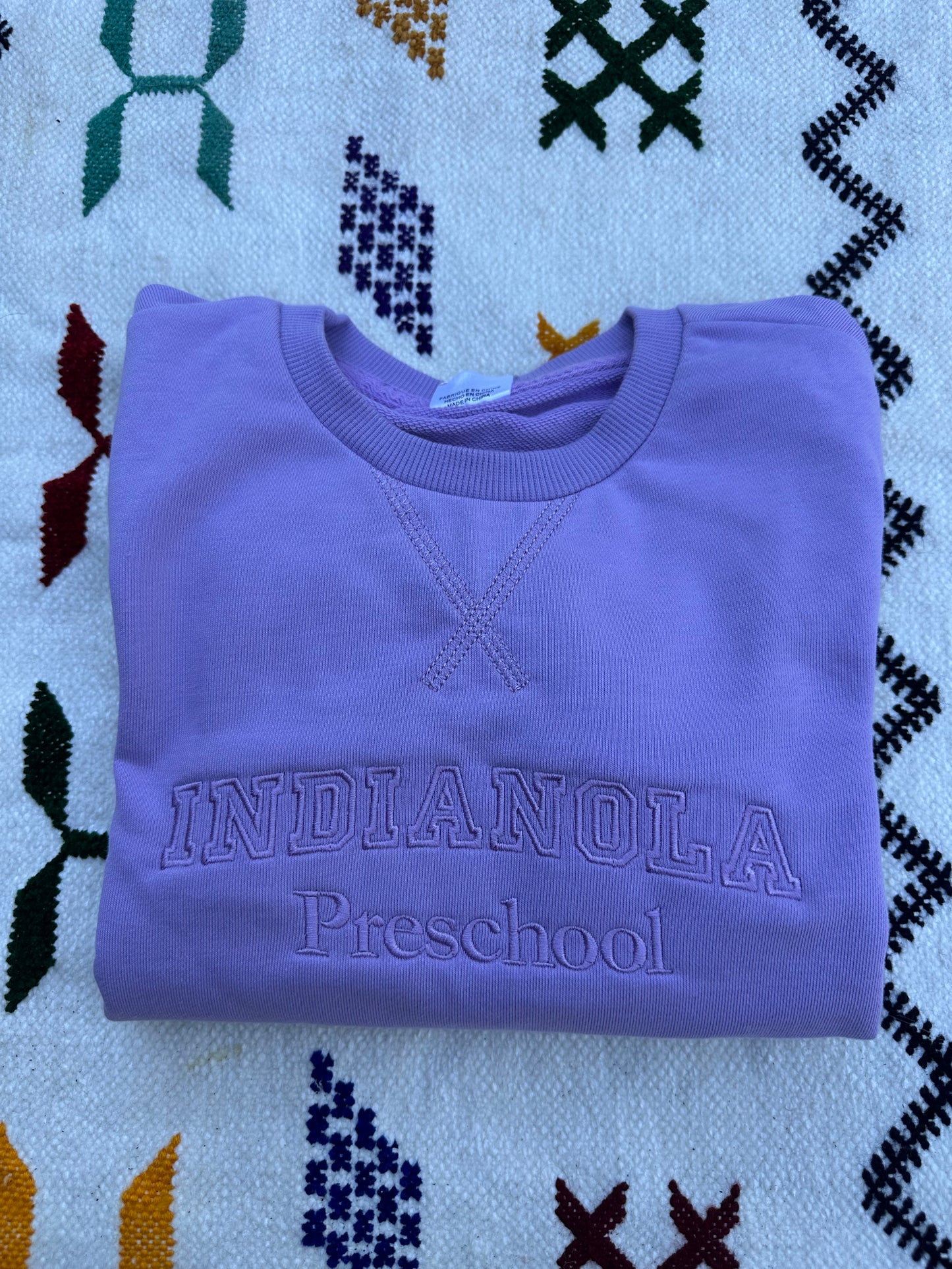 Folded purple sweatshirt with 'Indianola Preschool' text on a patterned fabric background