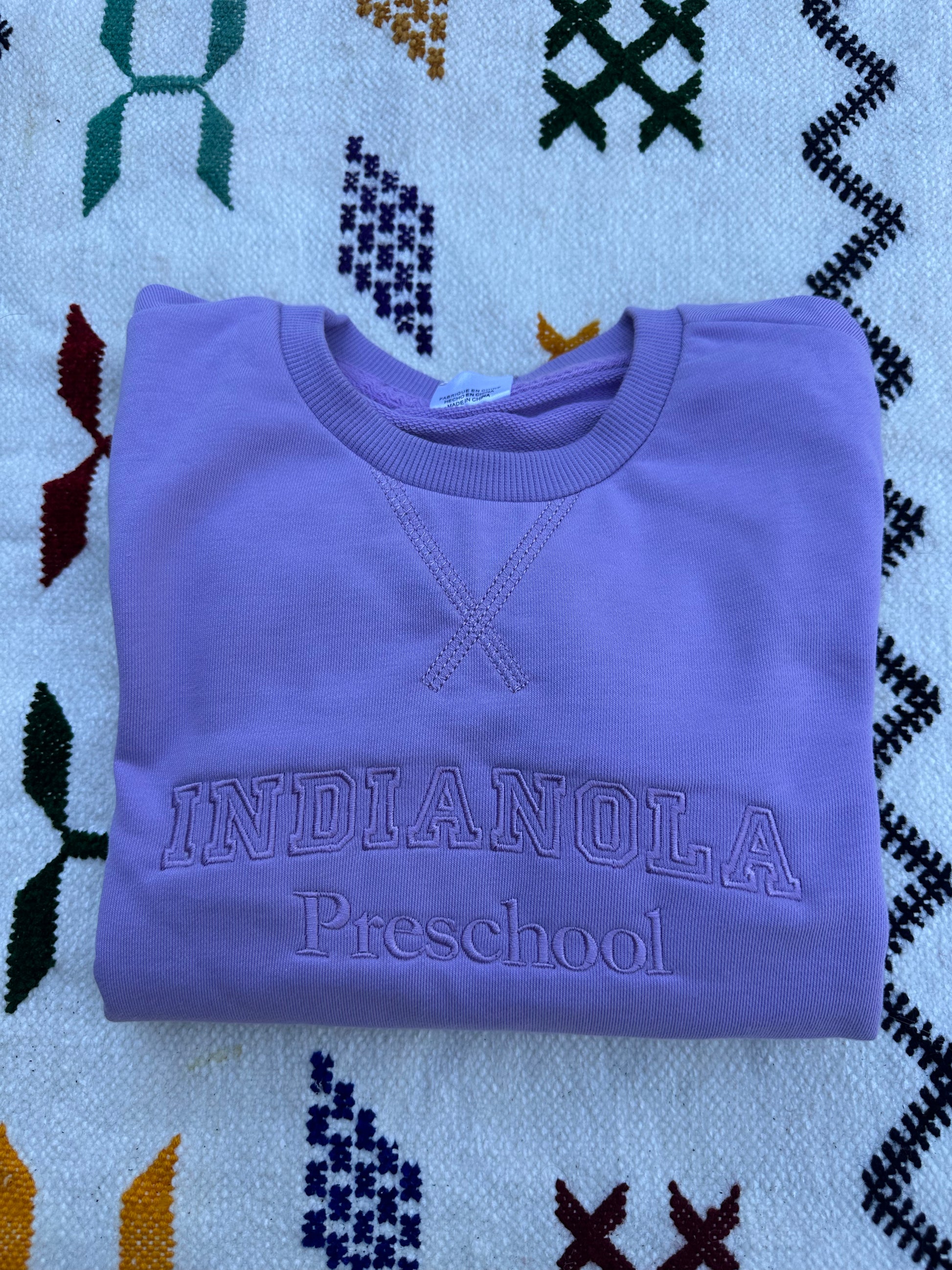 Folded purple sweatshirt with 'Indianola Preschool' text on a patterned fabric background